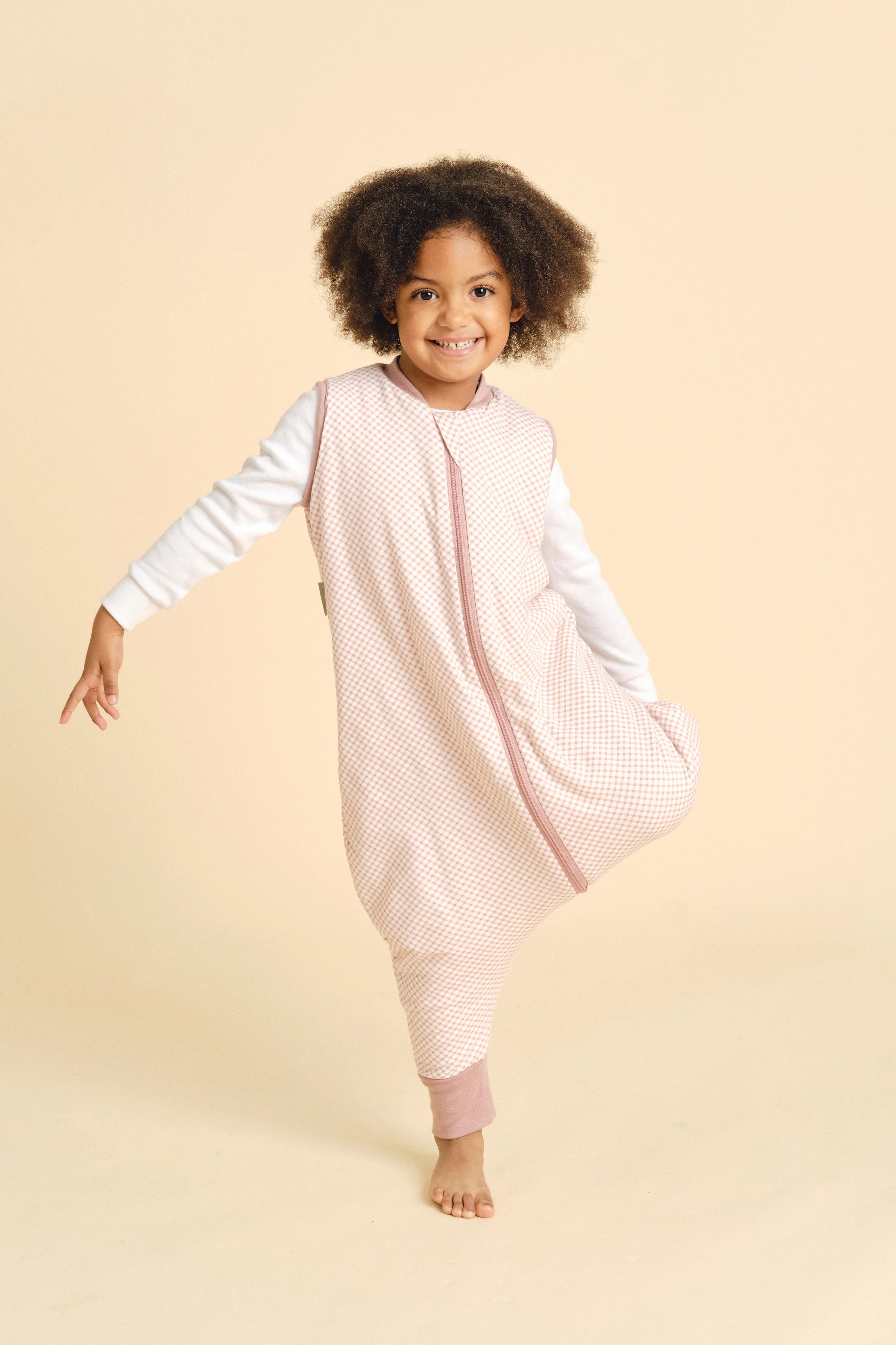 Criança mais velha sorridente, com cabelo encaracolado, a equilibrar-se numa perna com o saco de dormir MolisandCo TOG 1.0 VICHY PINK.