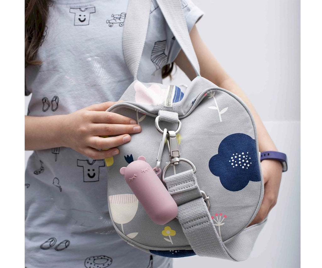 Criança a segurar uma mala cinzenta com padrão floral, onde está pendurado um estojo Keepie Palhinha de silicone rosa com mosquetão.