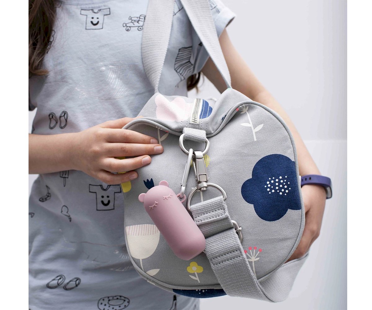 Criança a segurar uma mala cinzenta com padrão floral, onde está pendurado um estojo Keepie Palhinha de silicone rosa com mosquetão.