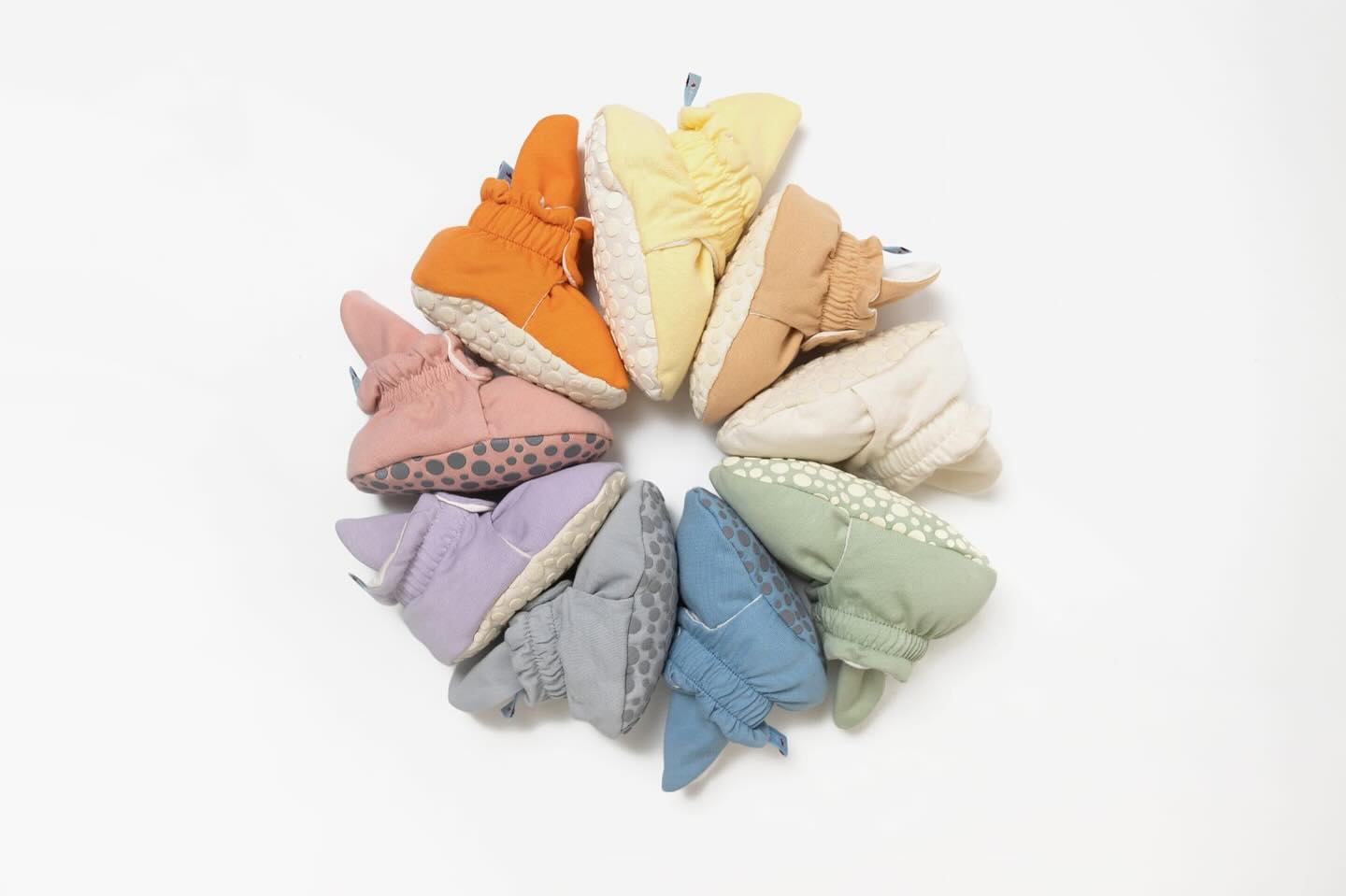 Conjunto de pantufas botinhas para bebé organizadas em círculo, exibindo várias cores pastel e tons suaves, incluindo laranja, amarelo, verde, azul, lilás e bege.
