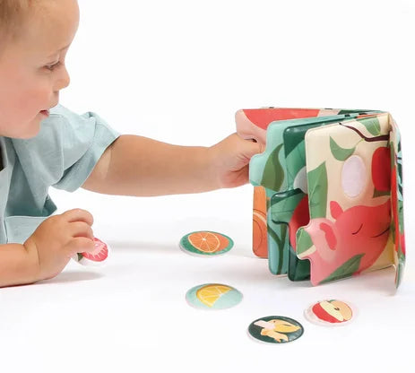 Mão de uma criança a interagir com o Ludi Livro de cheiros - Frutas, colocando uma peça de fruta no livro enquanto outras estão espalhadas.