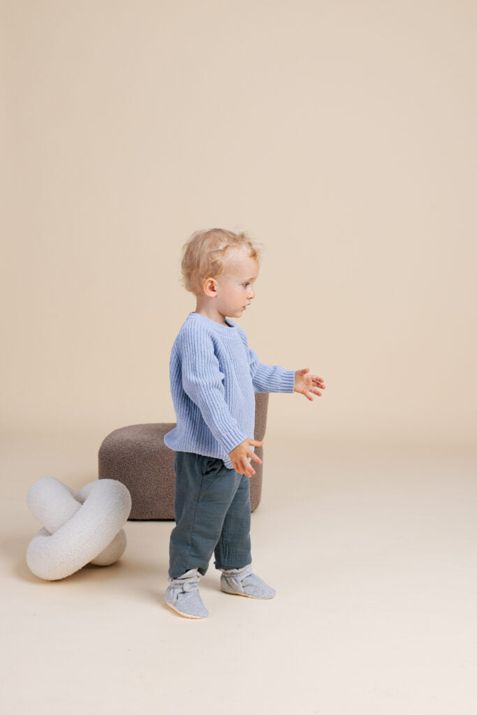 Bebé de cabelo loiro a dar os primeiros passos, usando as pantufas botinhas Zás Trás Mid Season Silver Pavlova, camisola azul e calças cinza, em fundo bege.