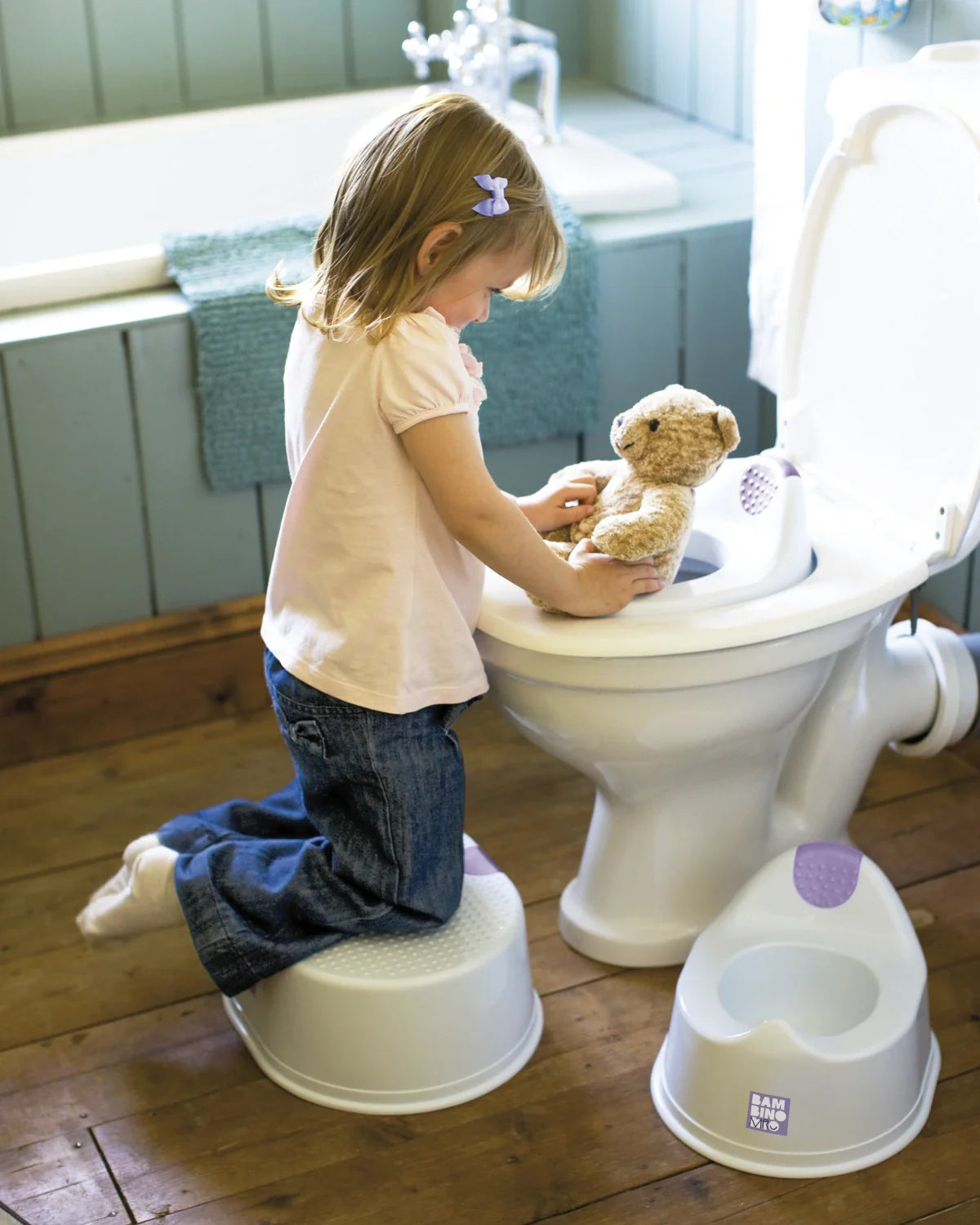 Menina a usar o degrau da Bambino Mio numa casa de banho, com o bacio Bambino Mio Pote no chão ao lado, mostrando o conjunto de desfralde.
