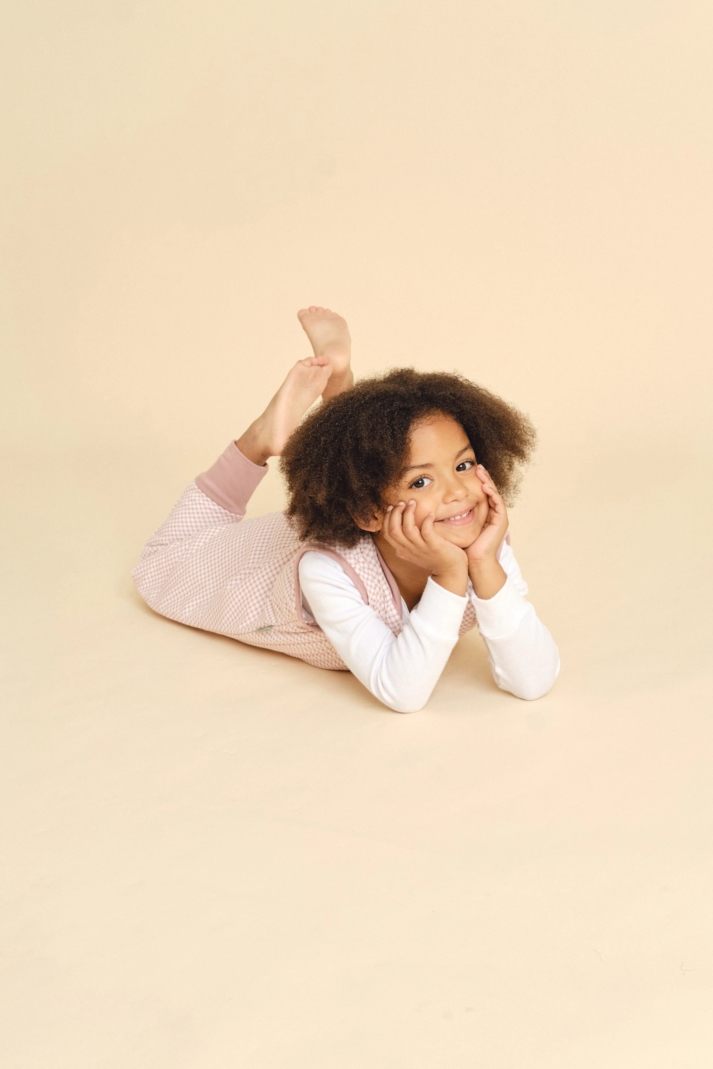 Criança mais velha sorridente, com cabelo encaracolado, deitada de bruços e com as mãos no queixo, a usar o saco de dormir MolisandCo TOG 1.0 VICHY PINK.