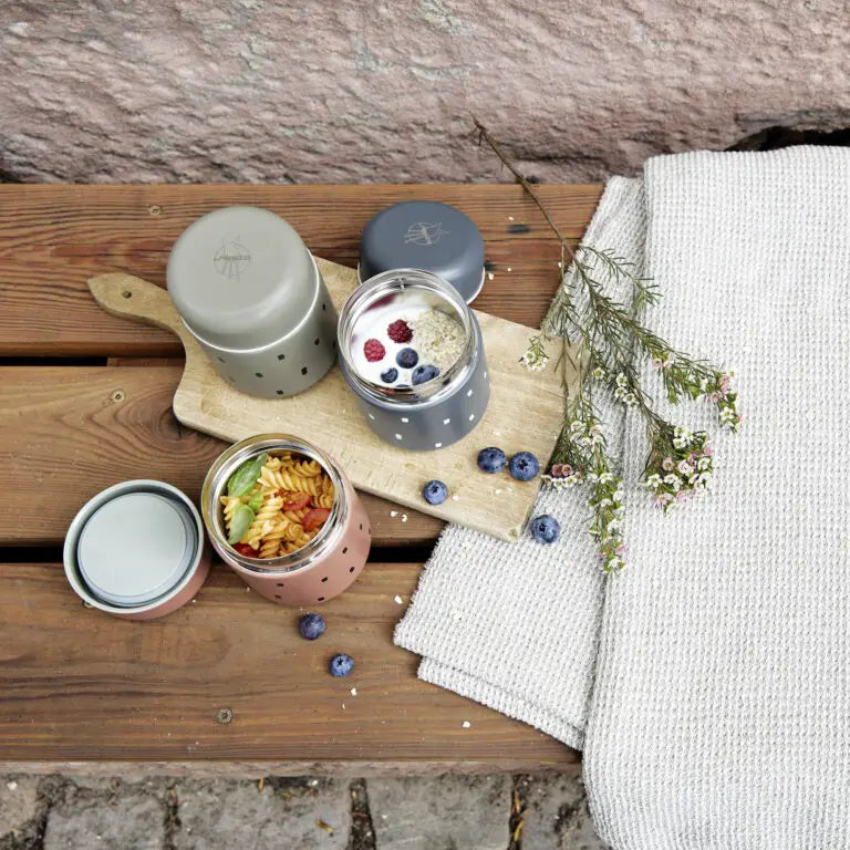 Dois termos Lassig de 350ml, um caramelo com massa e outro cinzento com iogurte e frutos vermelhos, sobre uma tábua de madeira rústica, com mirtilos e flores.
