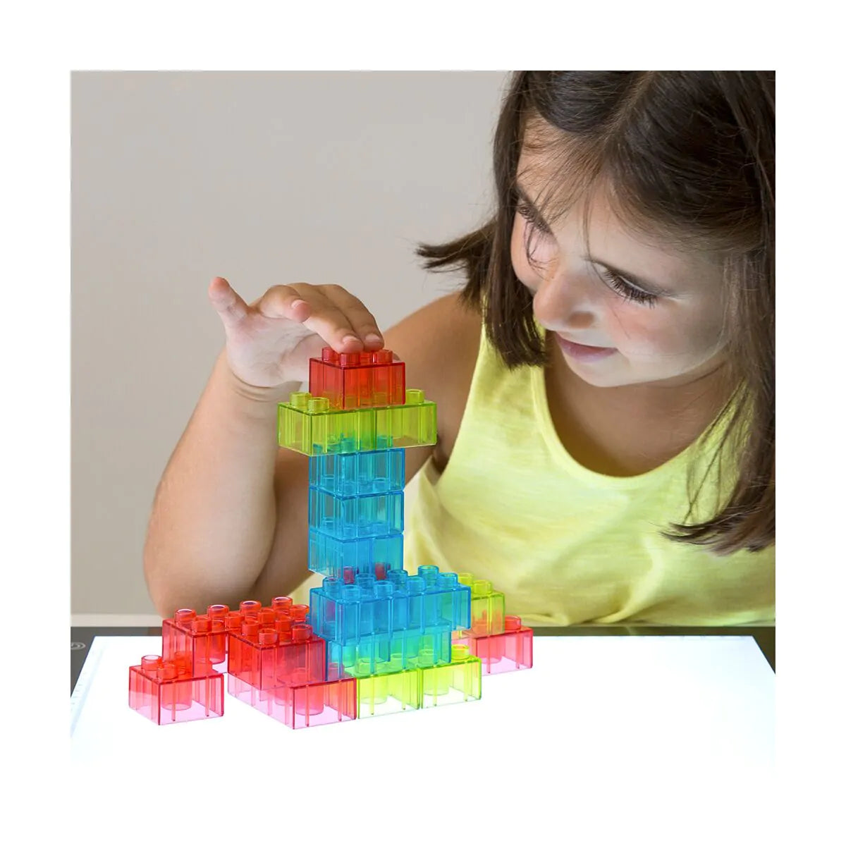 Uma criança a brincar com os blocos translúcidos Miniland numa mesa de luz, criando uma torre colorida.