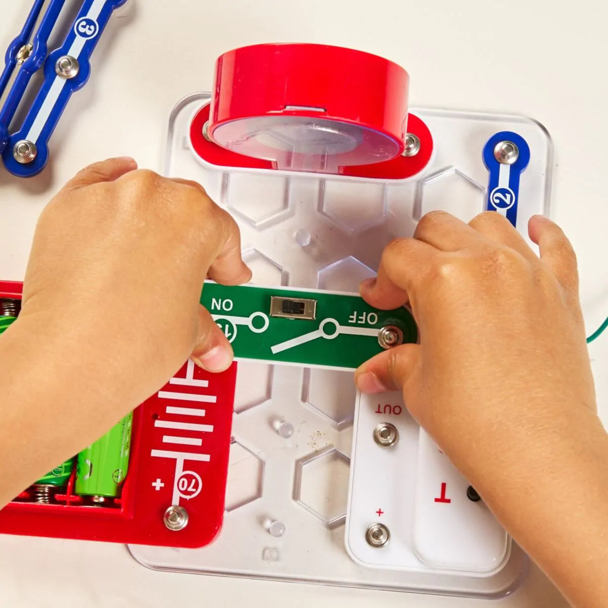 Mãos de criança a conectar um interruptor verde ao kit Miniland Electrofun: Tune In Radio, focando-se na placa transparente e nos componentes coloridos.