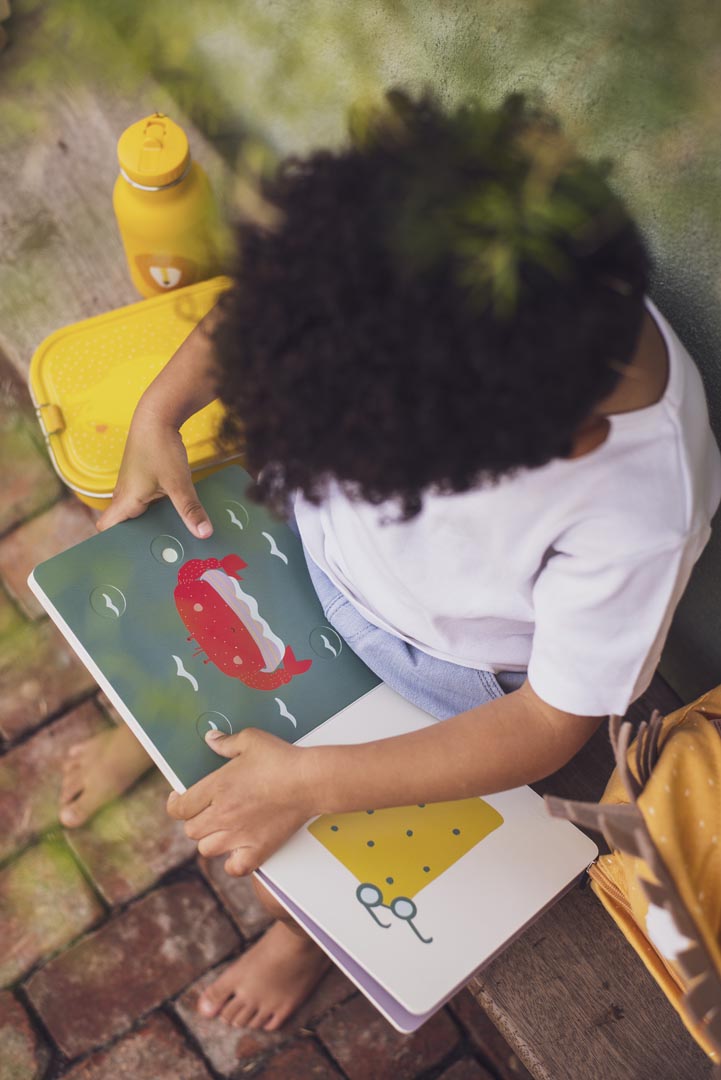 Vista superior de uma criança a folhear o livro Trixie, mostrando a página de uma baleia vermelha e a página seguinte com um desenho amarelo.