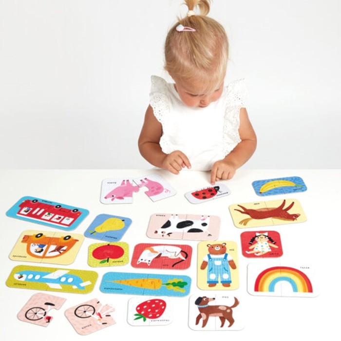 Uma menina pequena sentada a uma mesa a brincar com as peças espalhadas do puzzle Banana Panda First Words, concentrada a encontrar os pares.