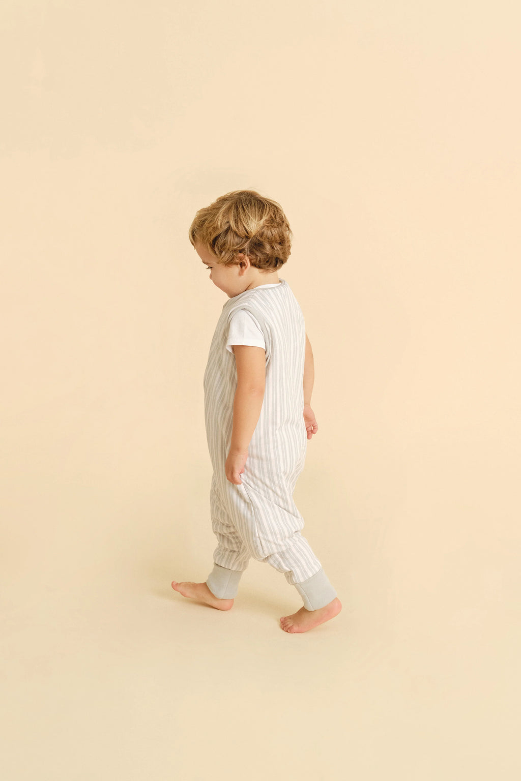 Criança loira a andar, de lado, a usar o saco de dormir MolisandCo TOG 1.0 Dawn Stripes com pés e meias, mostrando os pés descalços.