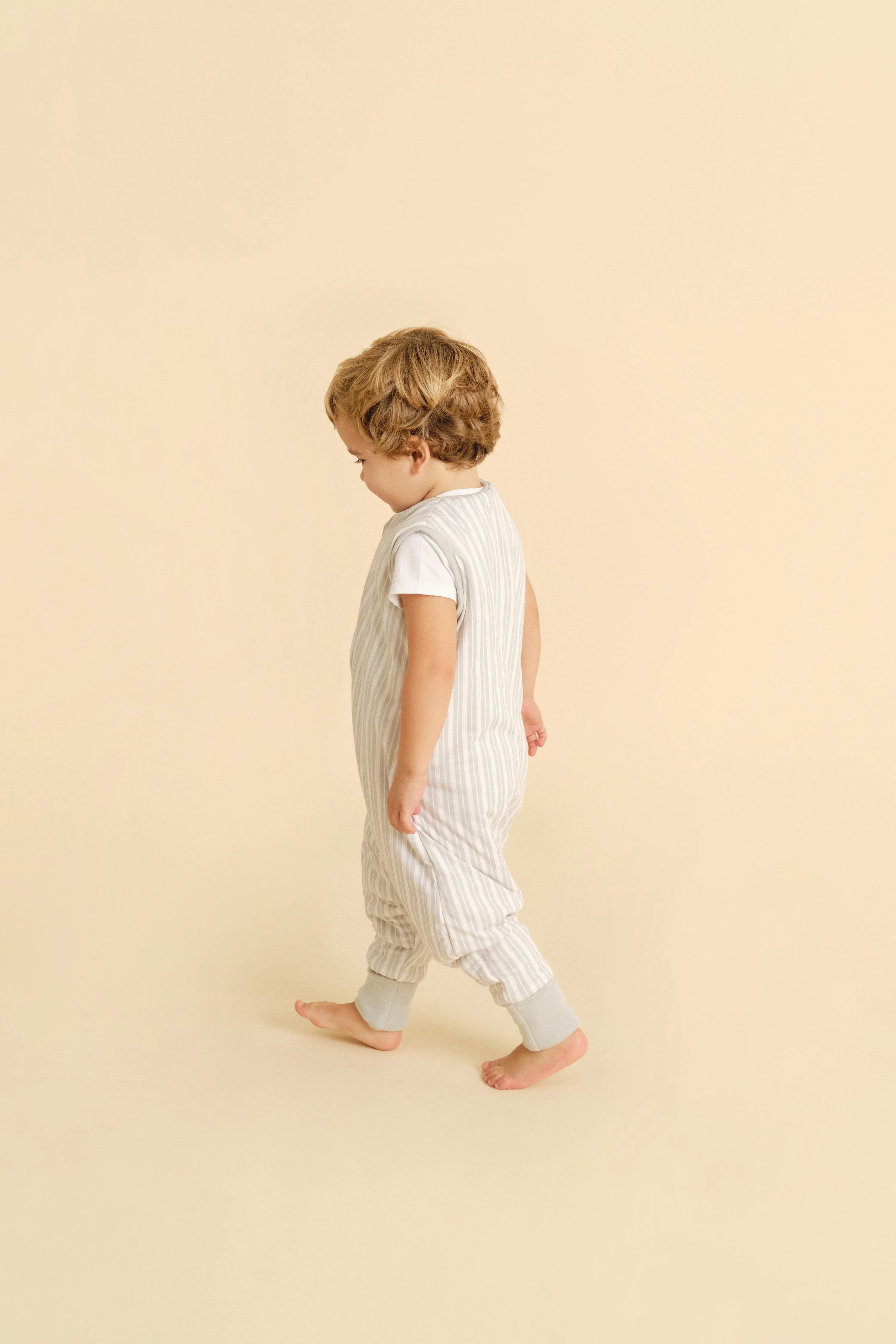 Criança loira a andar, de lado, a usar o saco de dormir MolisandCo TOG 1.0 Dawn Stripes com pés e meias, mostrando os pés descalços.