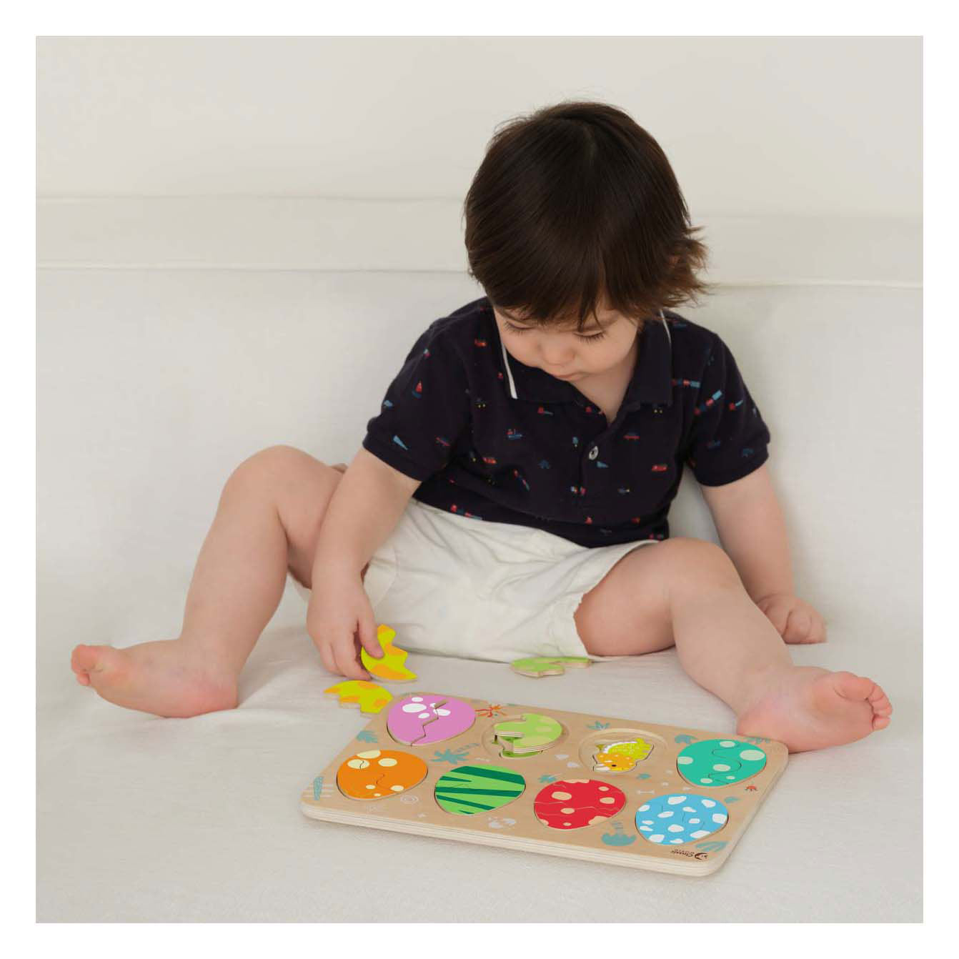 Uma criança pequena sentada a brincar com o Classic World Puzzle Encaixe Ovos de Dinossauro, concentrada a colocar uma peça no tabuleiro.