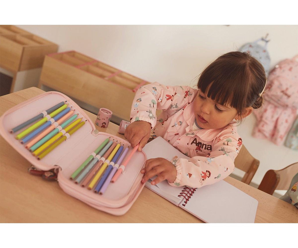Criança a escolher um lápis de cor de um estojo rosa, sentada numa secretária com um Caderno Escolar Tutete aberto.