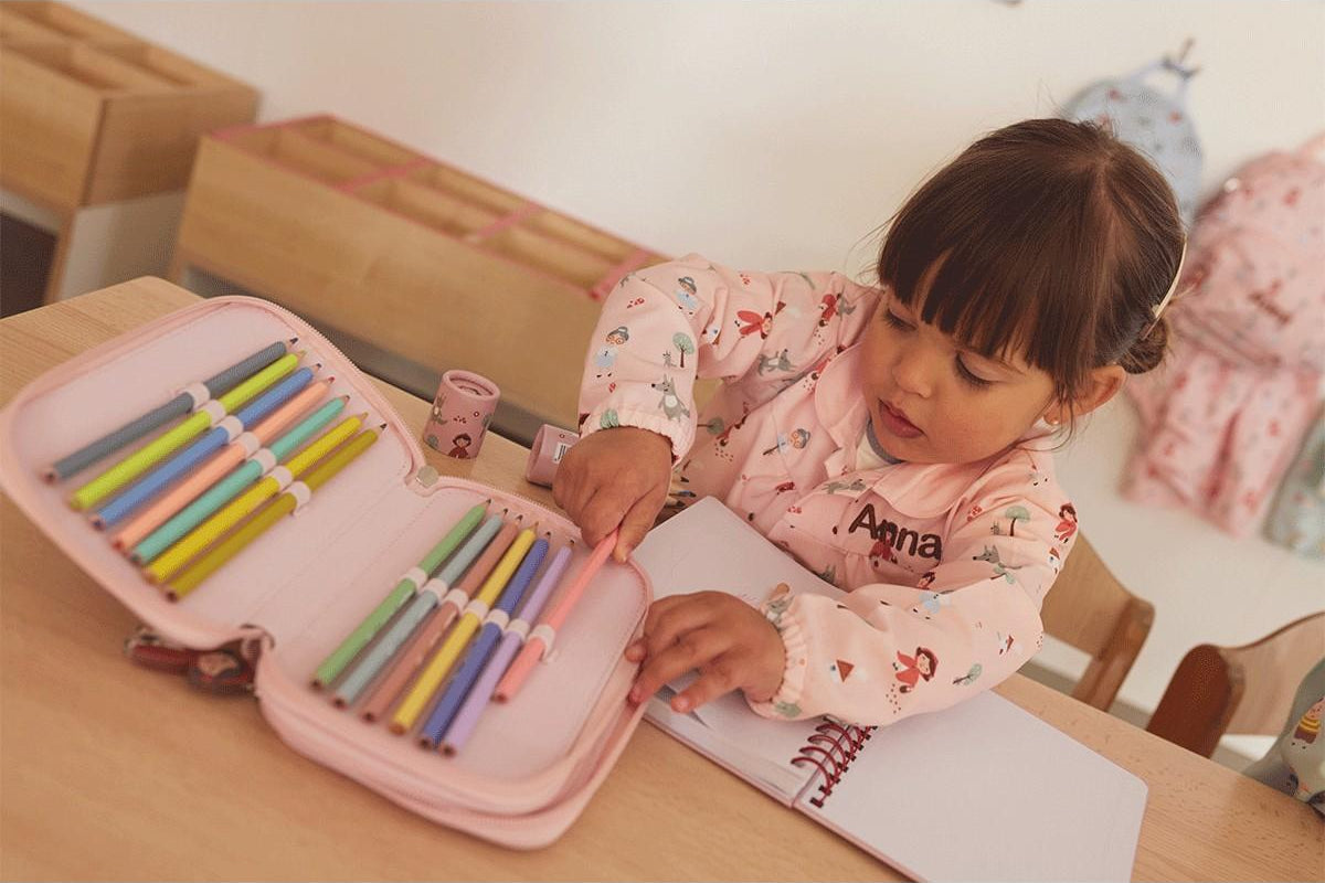 Criança a escolher um lápis de cor de um estojo rosa, sentada numa secretária com um Caderno Escolar Tutete aberto.