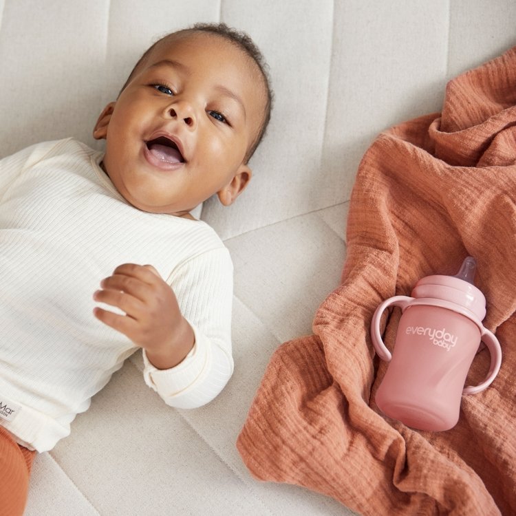 Bebé a sorrir deitado ao lado de um pano cor de terracota e um biberão rosa da Everyday Baby.