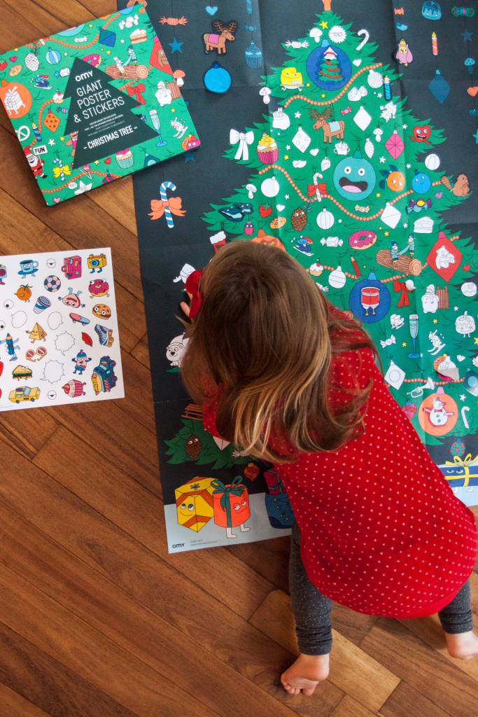 Uma criança a decorar um poster gigante de árvore de Natal com autocolantes, com a embalagem e uma folha de autocolantes ao lado.