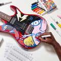 Pessoa a pintar uma guitarra elétrica vermelha com um marcador acrílico Carioca Plus, mostrando a versatilidade do produto em superfícies diversas.