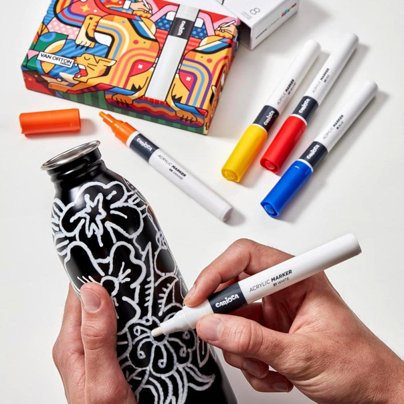 Mãos a decorar uma garrafa preta com um marcador acrílico Carioca Plus branco, rodeada por outros marcadores coloridos e a caixa.