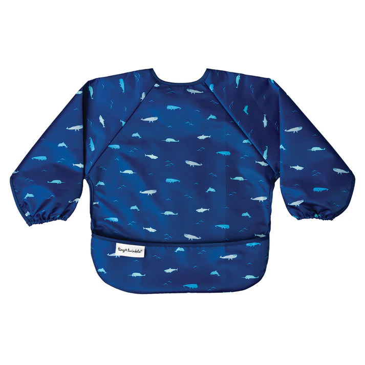 Babete impermeável Tiny Twinkle Oceano Life com mangas compridas e padrão de baleias e ondas em azul, visto em flat lay.