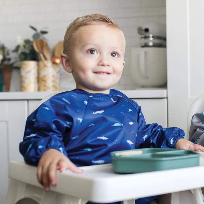 Criança sorridente a usar o babete impermeável Tiny Twinkle Oceano Life com mangas, sentada numa cadeira de refeição com um recipiente de comida.