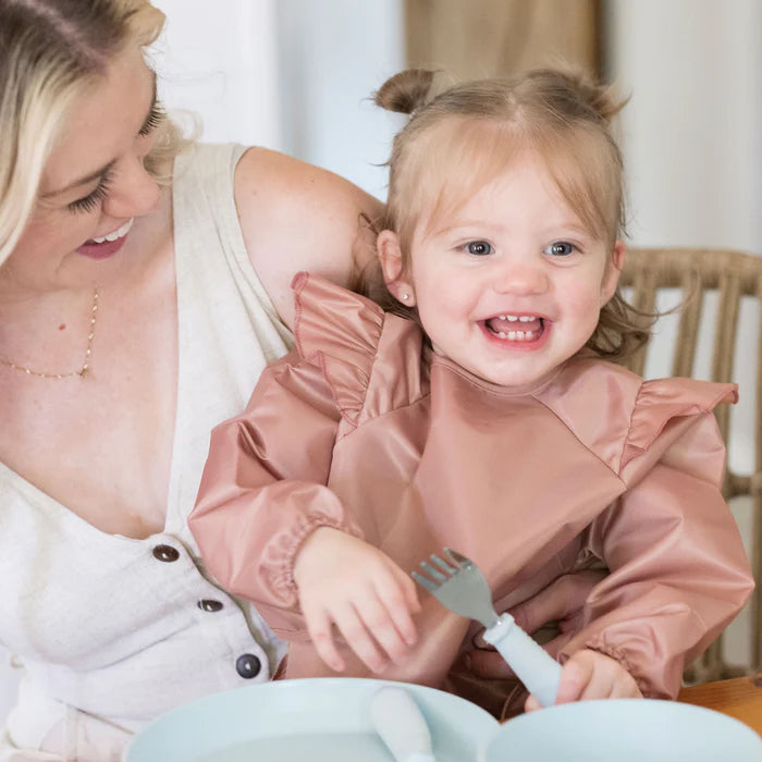 Criança sorridente com um babete de mangas compridas cor-de-rosa, sentada à mesa com um adulto, a segurar um garfo, ilustrando a utilização do babete.