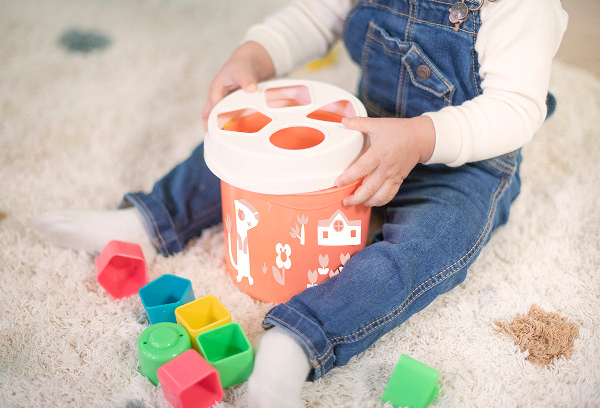 Mãos de bebé a segurar a tampa do Miniland Cubo com formas de encaixe laranja, com peças coloridas espalhadas ao redor.