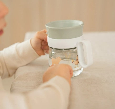 Criança a segurar o copo de aprendizagem Mepal 360º Little Farm pelas asas, pronta para beber de forma autónoma numa mesa.