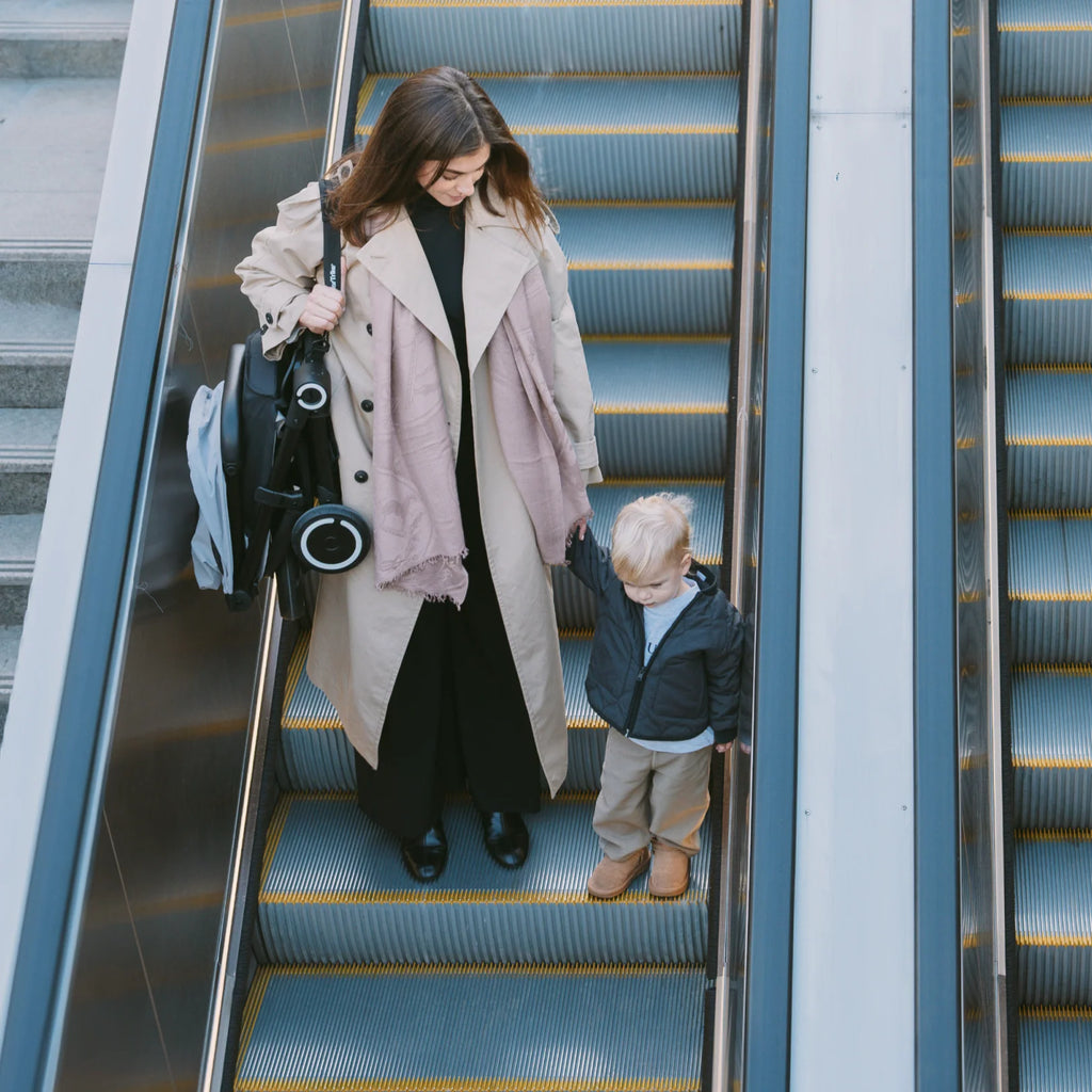 Mãe a transportar o Smartrike Traveler dobrado de forma compacta numa escada rolante, acompanhada pelo filho.