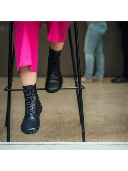 Detalhe de uma pessoa sentada num banco alto, vestindo calças cor-de-rosa vibrantes e as botas Angles Orpheus zip winter pretas.