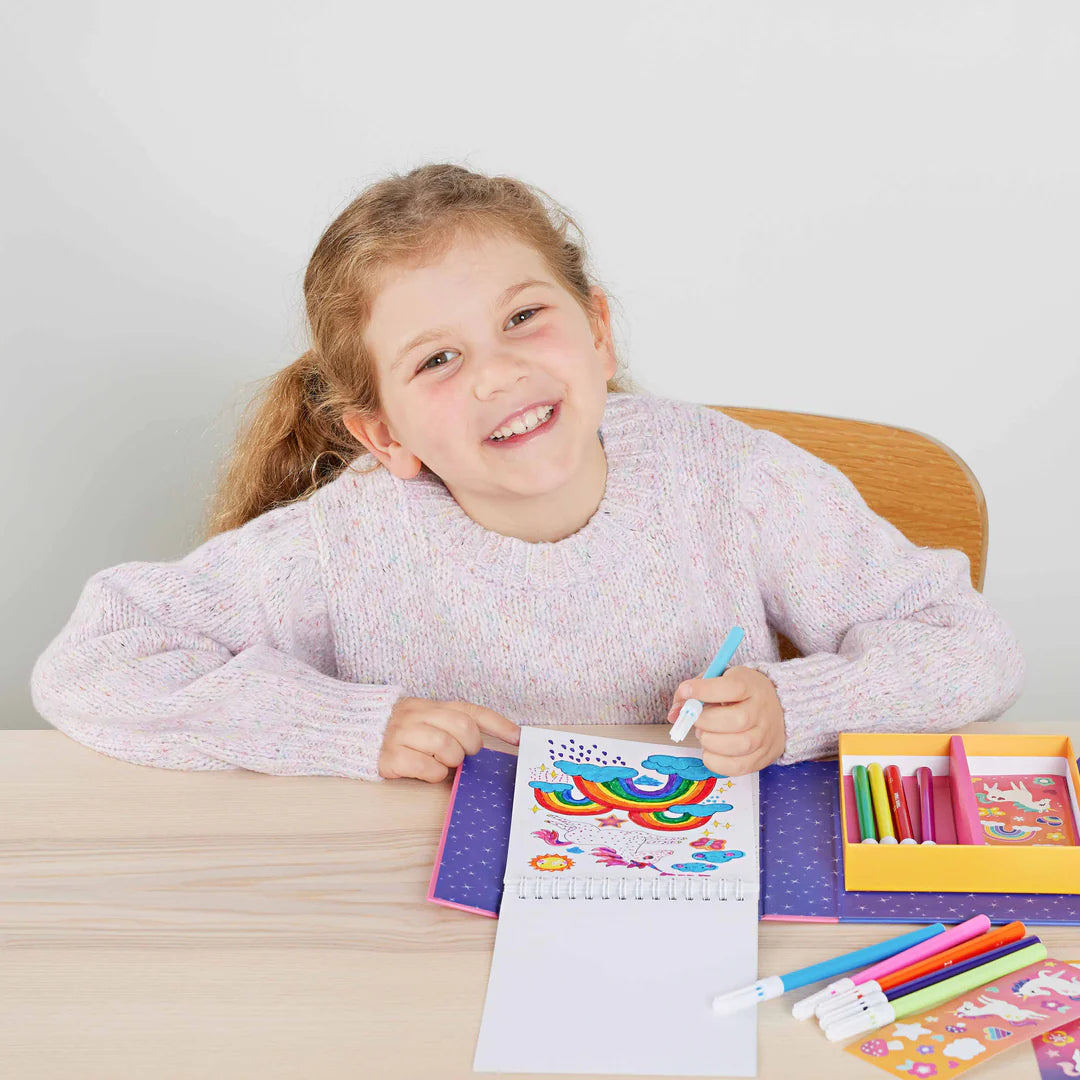 Criança sorridente a colorir uma ilustração de unicórnios e arco-íris num caderno do conjunto Tiger Tribe, com marcadores e a caixa aberta na mesa.
