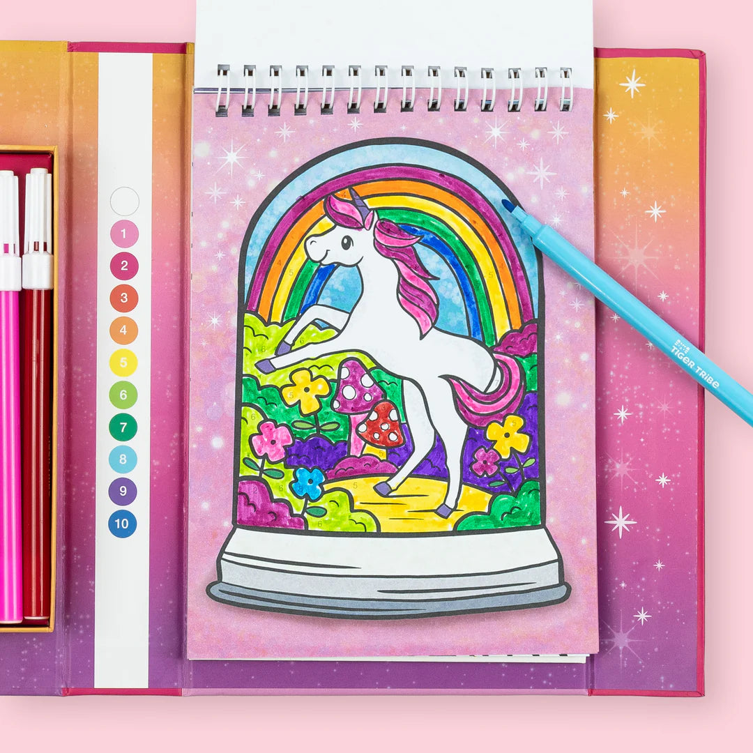 Caderno do conjunto Tiger Tribe aberto, mostrando uma ilustração de unicórnio dentro de um globo de neve para colorir por números, com um marcador azul pousado.