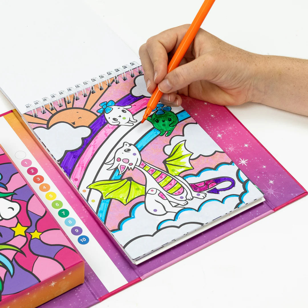 Mão de uma pessoa a colorir uma ilustração de um dragão e arco-íris com um marcador laranja no caderno do conjunto Tiger Tribe, com a paleta de cores numerada.