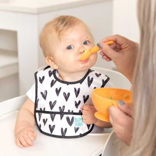 Bebé entre 3 e 9 meses sentado numa cadeira alta, a ser alimentado com uma colher por um adulto, usando o babete de corações.