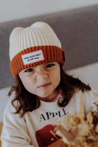 Menina com cabelo castanho a usar o gorro Hello Hossy Grandma Choco, com expressão divertida e camisola com desenho de maçã.