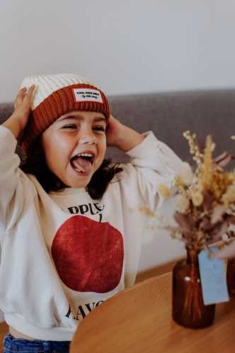 Menina a rir com o gorro Hello Hossy Grandma Choco na cabeça, mostrando a sua alegria e o estilo do acessório.