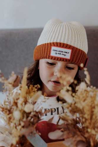 Menina com o gorro Hello Hossy Grandma Choco, olhando para baixo com curiosidade, em ambiente acolhedor.