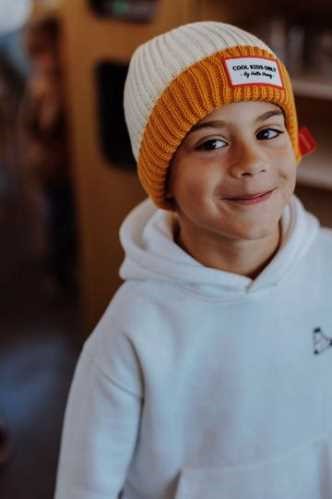 Criança sorridente a usar o gorro Hello Hossy Grandma Curry, com a parte superior creme e a dobra mostarda, sobre uma camisola branca.