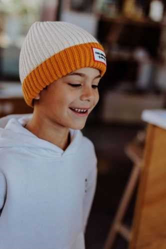 Criança a usar o gorro Hello Hossy Grandma Curry, com a parte superior creme e a dobra mostarda, olhando para baixo e sorrindo.