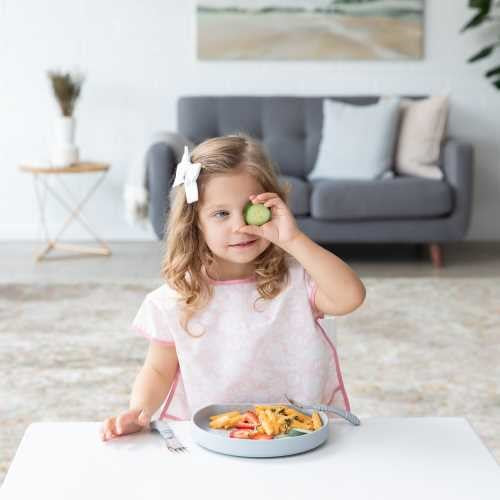 Criança pequena sorridente, vestindo o babete de mangas rosa, a segurar um pedaço de pepino com os dedos junto ao olho enquanto está sentada à mesa com um prato de comida.