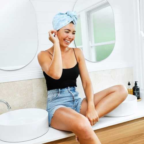 Mulher sorridente sentada na bancada da casa de banho, a usar a touca de secagem rápida Dock and Bay com padrão azul-claro.