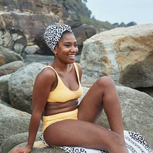 Mulher sorridente sentada em rochas na praia, vestindo um fato de banho amarelo e a touca de secagem rápida Dock and Bay.