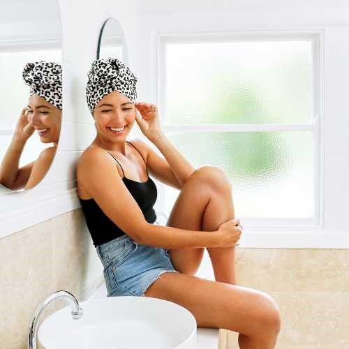 Mulher feliz sentada na bancada da casa de banho, a usar a touca de secagem rápida Dock and Bay com padrão de leopardo.