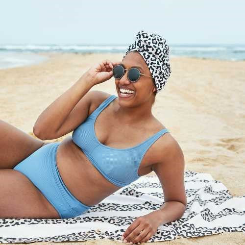 Mulher deitada numa toalha na praia, a usar óculos de sol e a touca de secagem rápida Dock and Bay com padrão de leopardo.