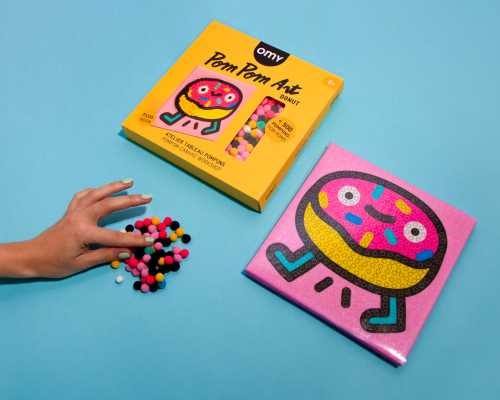 Kit OMY Pintura com Pompons Donut: tela, caixa e mão a pegar em pompons coloridos numa superfície azul.