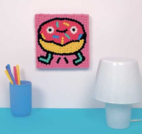 Kit OMY Pintura com Pompons Donut: donut de pompons colorido pendurado numa parede branca, junto a uma secretária.