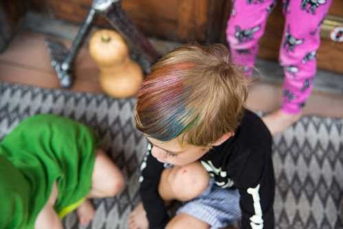 Criança com cabelo loiro curto, com madeixas coloridas de azul e vermelho, sentada num tapete.