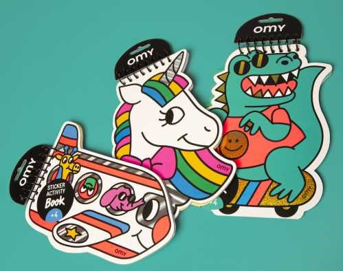 Livros de autocolantes OMY em forma de unicórnio e dinossauro, com embalagem preta, sobre fundo azul-petróleo.