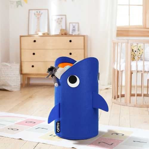 OMY Cesto de Arrumação em forma de tubarão azul, cheio de brinquedos, posicionado num quarto infantil com um berço e cómoda ao fundo.