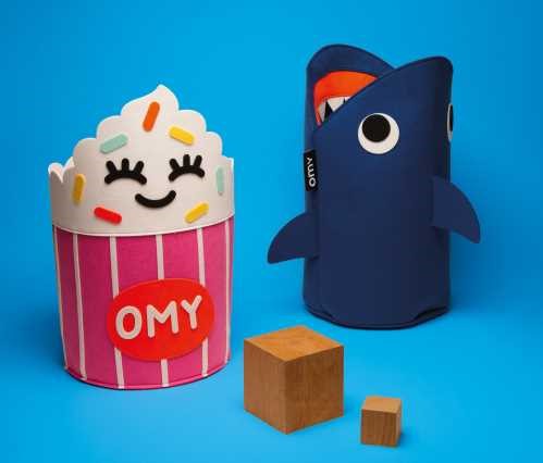 OMY Cesto de Arrumação em forma de tubarão azul ao lado de um cesto em forma de cupcake rosa, ambos sobre um fundo azul liso, com dois cubos de madeira em primeiro plano.