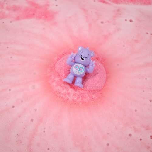 Figura roxa do Ursinho Carinhoso com símbolos de pirulito ao centro de uma bomba de banho rosa efervescente.